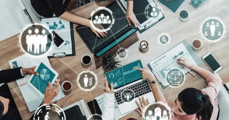 Overhead view of a team collaborating around a table with laptops, tablets, and printed charts, including a document labeled “Project Plan Year.” Semi-transparent icons of people, networks, and analytics overlays connect across the scene, suggesting hiring, workforce strategy, and data-driven team building.