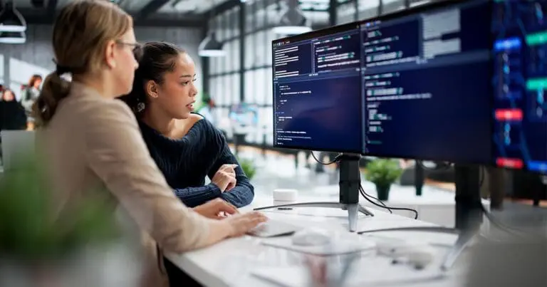 Two colleagues sit at a desk in a modern office, focused on a large monitor displaying lines of code. One points toward the screen while the other types, suggesting collaboration on a software or programming task.