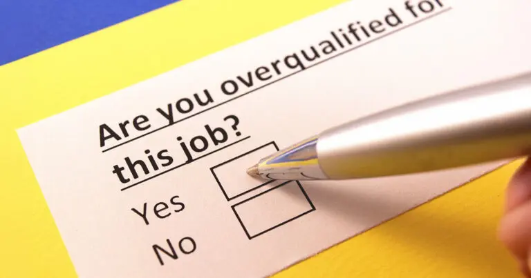 Close-up of a printed form asking “Are you overqualified for this job?” with “Yes” and “No” checkboxes, as a pen hovers over one option. The document rests on a bright yellow background, emphasizing the decision point.