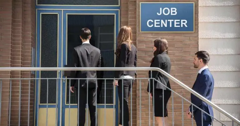 Four professionally dressed adults stand in line outside a building labeled “JOB CENTER,” facing a closed door as they wait to enter. A metal railing runs along the steps, and the scene conveys a formal, subdued mood of job seeking and uncertainty.