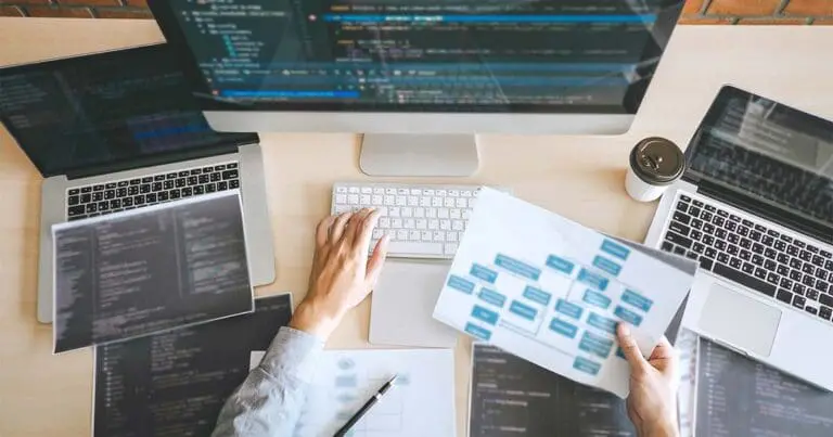 Overhead view of a desk with multiple laptops and a large monitor displaying lines of code, while a person uses a keyboard and holds a printed workflow diagram. The scene conveys a busy development environment with overlapping screens and tools, suggesting complexity and coordination in web development work.
