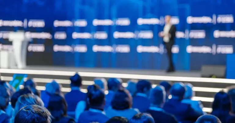 Blurred audience seated in front of a large conference stage with a speaker presenting against a blue backdrop. The image conveys a corporate technology event or keynote presentation to represent trustworthy ai thought leaders worth following.