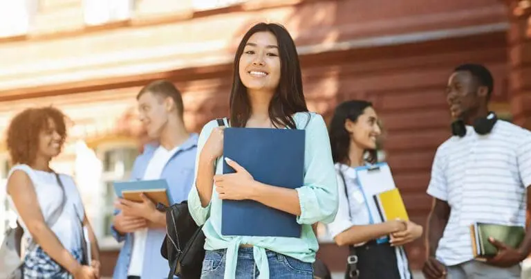 Smiling college student stands in the foreground holding a notebook and backpack, with a diverse group of classmates chatting behind her outside a campus building. Warm sunlight and casual poses create an optimistic, forward-looking tone to represent Job Search Tips for New Graduates.