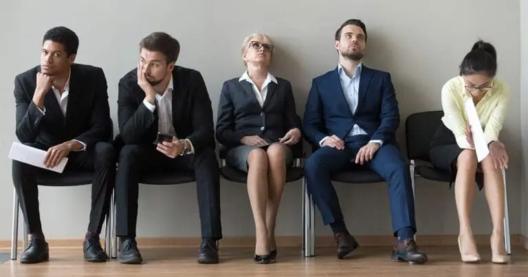 Five job candidates in business attire sit in a row against a plain wall, each displaying signs of boredom or frustration while waiting, including slouching, looking away, and checking a phone. One woman on the far right leans forward reading a document, contrasting with others who appear disengaged and restless all to symbolize reasons candidates drop out of hiring processes.