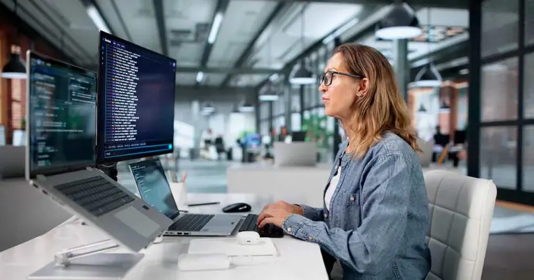 A software professional works at a multi-screen workstation displaying lines of code in a modern open office. The scene conveys a collaborative, technology-driven work environment, reflecting how coding practices and technical evaluation are evolving in the hiring process.