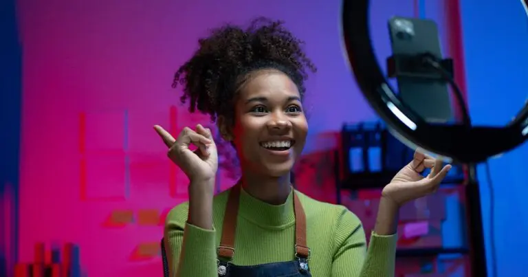 A content creator smiles and gestures while recording a video on a smartphone mounted inside a ring light, with colorful pink and blue studio lighting in the background. The image illustrates the growing role of influencer content, social commerce, and creator-driven marketing in digital marketing careers and hiring.