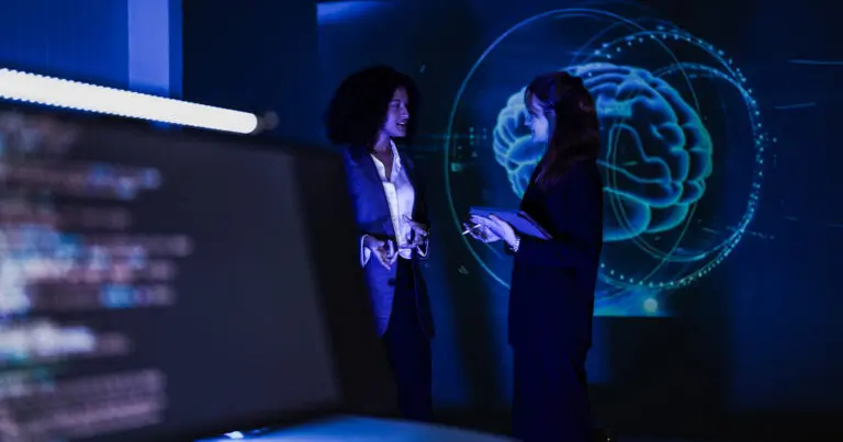 Two professionals discuss AI research in a dark, blue-lit lab, with a glowing digital brain visualization projected behind them and blurred code on a foreground screen.