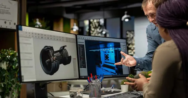 Two colleagues review technical designs on dual computer monitors in an office, with one person gesturing toward a 3D model and interface displayed on the screens while explaining details to the other. The collaborative setting and focus on digital prototypes convey a discussion around engineering, product development, or design work.