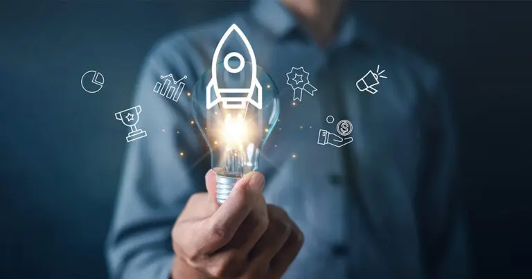 A person holds a glowing light bulb with a rocket icon inside, surrounded by line-drawn business symbols including growth charts, a trophy, an award ribbon, marketing, and funding icons. The image conveys innovation, startup formation, and the early-stage momentum associated with building teams and launching new ideas from zero to one.