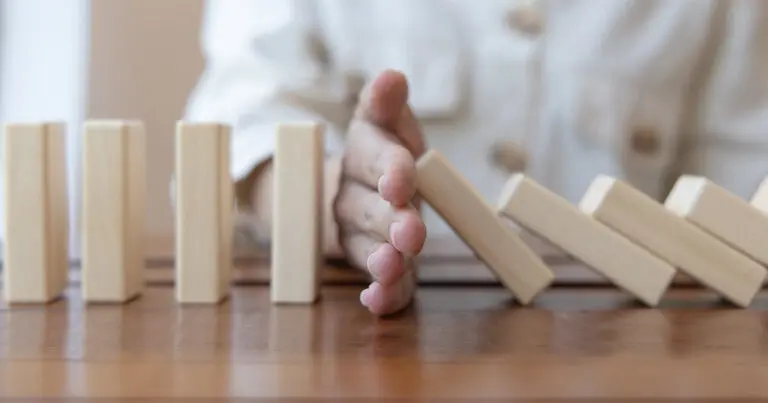 A hand reaches between upright and falling wooden domino blocks on a tabletop, stopping the chain reaction before it spreads further. The scene conveys prevention, risk management, or interrupting a cascading problem.