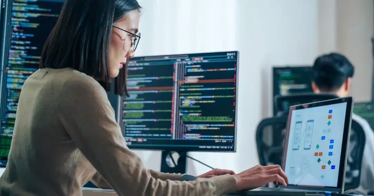 A software developer works at a desk with multiple screens displaying code while using a laptop that shows a mobile app interface and workflow diagram. Another developer is visible out of focus in the background, reinforcing a collaborative engineering environment and illustrating the topic of AI-assisted coding and changing developer hiring practices.