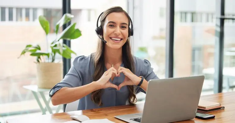 A smiling customer service employee wearing a headset sits at a desk with a laptop, forming a heart shape with their hands during a video call in a bright office. The warm, welcoming gesture reinforces the theme of employee engagement and positive workplace culture for an article about effective employee retention strategies to reduce turnover.