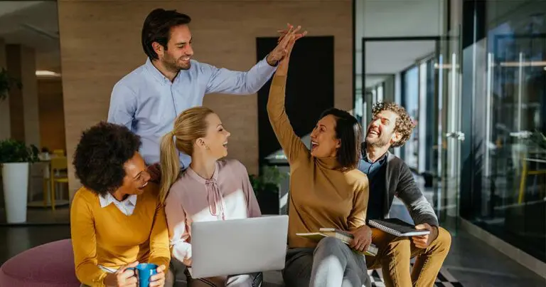 A group of five coworkers in a modern office smile and celebrate as two colleagues exchange a high-five while others look on. A laptop, notebooks, and coffee mug suggest a collaborative meeting, with natural light creating an upbeat, team-oriented atmosphere.