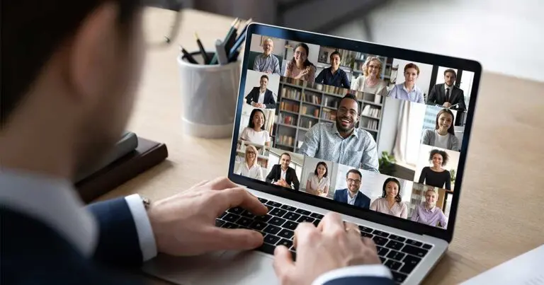 A person works at a laptop displaying a video conference with multiple participants arranged in a grid, suggesting a virtual team meeting or collaboration session. The scene illustrates remote work and highlights the continued use of online meetings by distributed employees and employers.