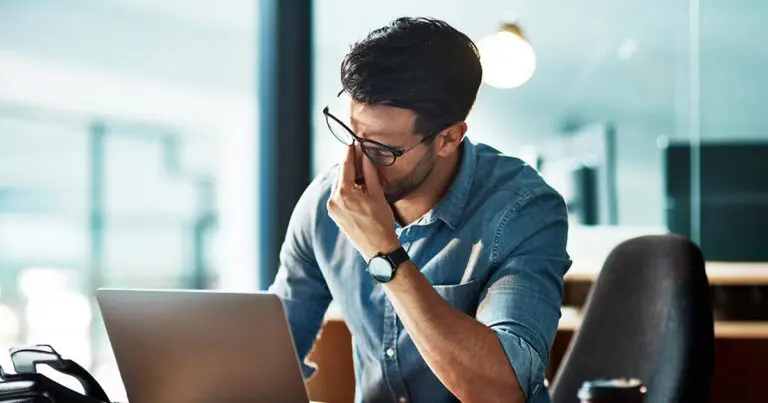A person wearing glasses sits at a desk in front of a laptop, pressing their fingers to the bridge of their nose in a gesture of stress or fatigue. The office setting and subdued posture convey the pressure and exhaustion associated with an overwhelming workload or burnout.