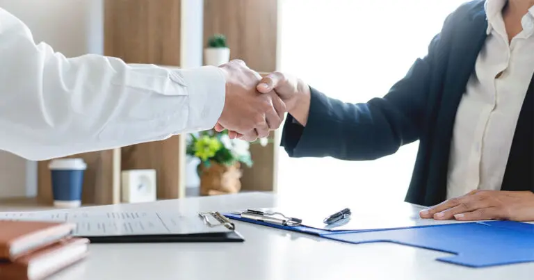 Two business professionals shake hands across a desk with a resume, clipboard, and signed documents in the foreground, symbolizing a hiring agreement. The close-up composition emphasizes recruitment, professional partnerships, and the business implications of specialized talent hiring, aligning with the theme of the hidden costs of DIY specialized talent recruiting.