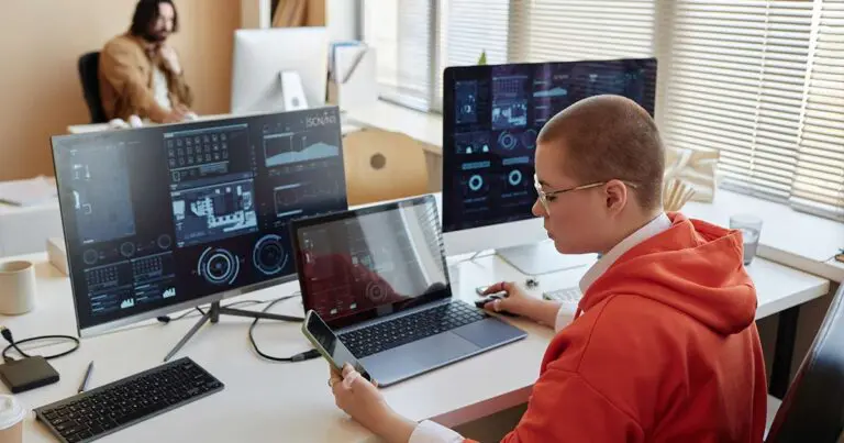 An employee in an orange hoodie works at a desk with multiple monitors displaying cybersecurity dashboards and analytics while holding a smartphone, with another colleague seated in the background of the office. The scene highlights the human and operational side of cybersecurity work, reflecting the staffing demands behind maintaining cyber resilience.