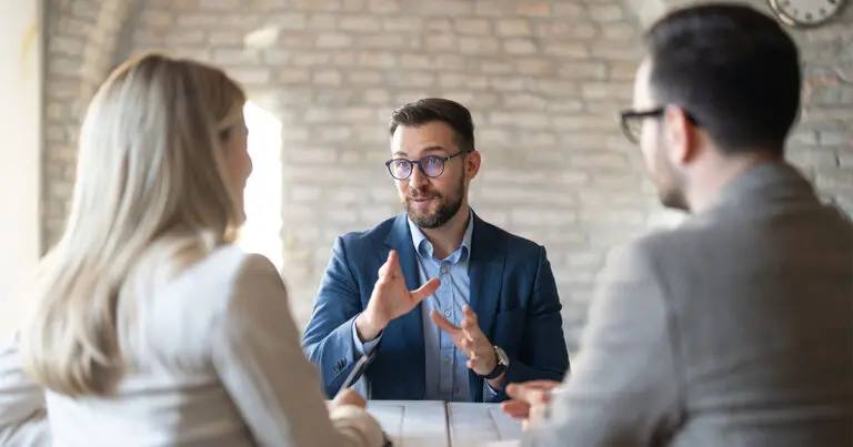 A recruiter in a blue blazer gestures while speaking to two hiring managers seated across a conference table in a modern office with a brick wall backdrop. The meeting conveys a collaborative discussion about hiring strategy before opening a new role.