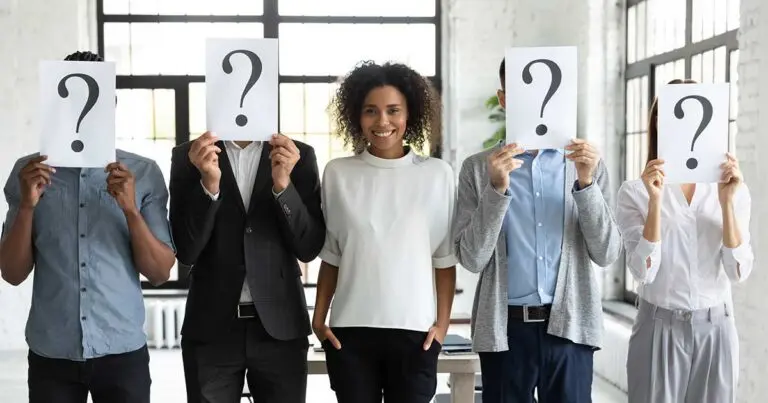 A diverse group of five job candidates stands in an office, with four holding sheets displaying large question marks that cover their faces while one smiling person remains visible in the center. The image conveys the challenge of identifying strong hiring decisions and choosing the right candidate, matching the theme of traits that lead to better hiring outcomes.