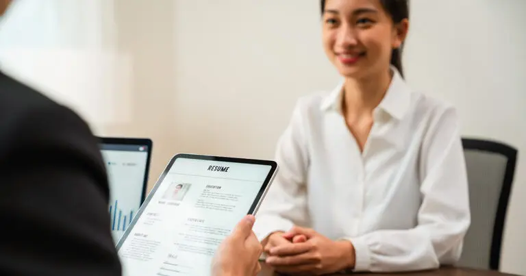 A job candidate sits across from an interviewer holding a tablet displaying a résumé during a formal interview in an office. The image illustrates how AI has made job interviews the weakest link in the hiring process by highlighting the traditional interview setting where candidates are evaluated face to face.