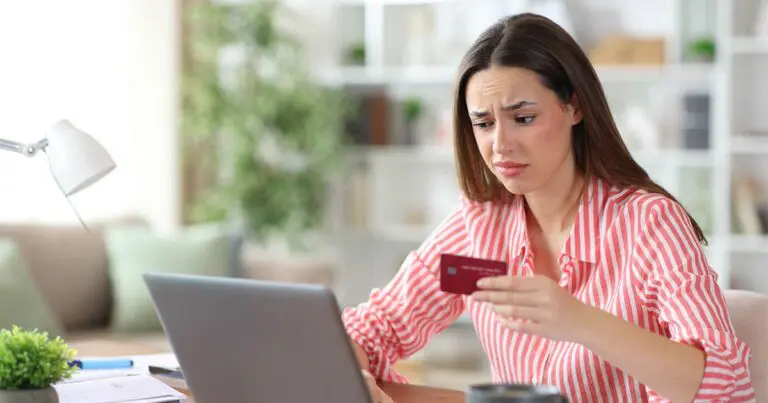 A concerned woman holds a credit card while looking at a laptop at a desk, her furrowed expression suggesting frustration or confusion.
