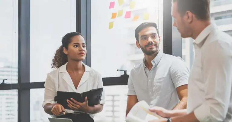 Three professionals stand together in a bright office during a meeting, with one person holding a notebook while another presents printed documents and the group listens attentively. Colorful sticky notes are posted on a window behind them, reinforcing the collaborative planning and decision-making theme of an article about questions hiring managers should ask before opening a new role.