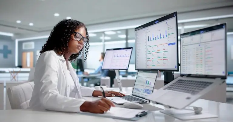 A person in a white lab coat works at a desk with multiple monitors displaying dashboards, charts, and system management interfaces while taking notes. The modern office setting and focus on monitoring data and digital systems reinforce the article’s theme of evaluating the skills and characteristics needed when hiring a system administrator.