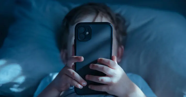 A child lies in bed holding a smartphone close to their face, illuminated by the screen’s blue light in a darkened room.