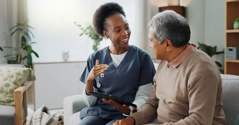 A home care professional in scrubs smiles and speaks with an older man while holding a tablet as they sit together on a couch, illustrating the growing role of caregivers and technology in aging at home.