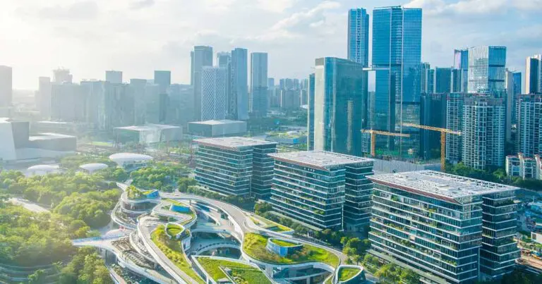 Aerial view of a modern urban business district with glass high-rises, construction cranes, landscaped office buildings, and sweeping green-roofed structures, illustrating the physical infrastructure behind the growing AI economy.