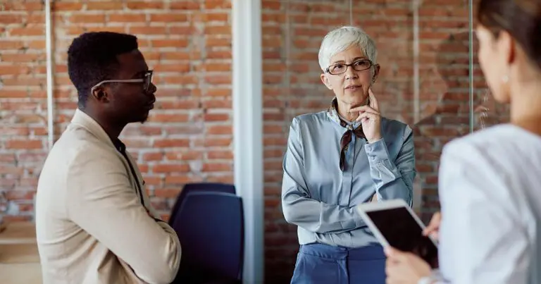 Three professionals hold a focused discussion in a modern office, with an older woman speaking thoughtfully as two colleagues listen. The scene reflects collaboration, leadership, and strategic business planning.