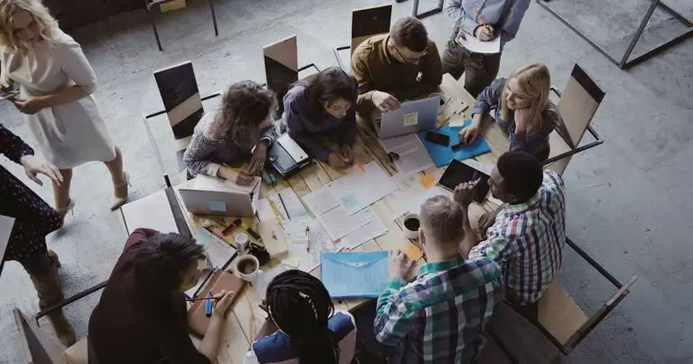 A diverse group of coworkers gathers around a large table covered with laptops, documents, notebooks, and coffee as they collaborate in an open office.