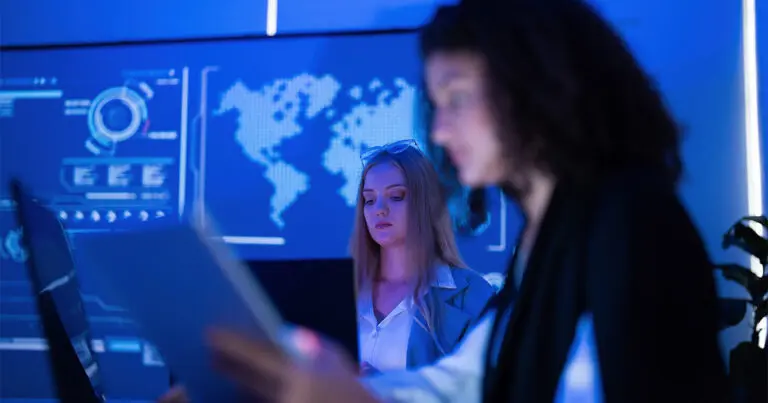 Two cybersecurity professionals work at computer monitors in a blue-lit operations center, with digital dashboards and a world map displayed behind them. The scene reflects the growing technical and staffing demands associated with AI, cloud adoption, and cybersecurity.