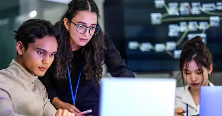 Three coworkers collaborate around laptops in an office, with one woman leaning over a seated colleague’s screen while another works nearby. A large display behind them shows a network of connected digital elements, reinforcing the theme of human collaboration and judgment in an AI-enabled workplace.