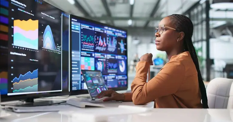 A business analyst wearing glasses and an orange blazer studies multiple computer monitors displaying colorful data visualizations, charts, and analytics dashboards in a modern office. A laptop with additional data graphics sits on the desk in front of her, highlighting the role of data analysis in digital transformation.