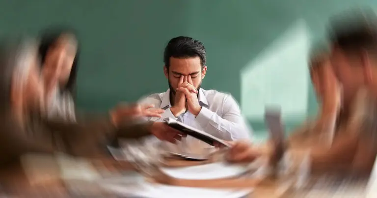 A stressed employee sits at a meeting table with his eyes closed and hands clasped against his face, surrounded by blurred coworkers handling documents. The motion blur emphasizes the hectic workplace environment, illustrating the pressure of being short-staffed or under-resourced.