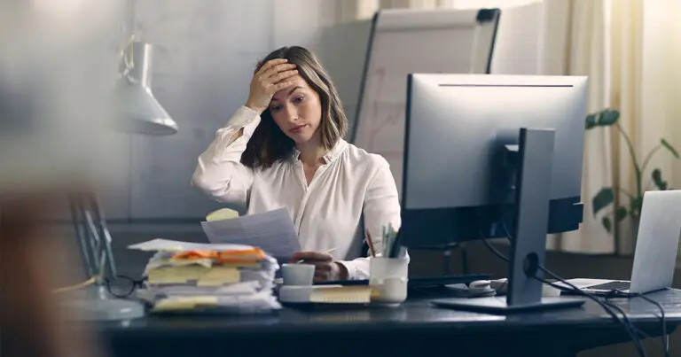 A stressed office worker sits at a cluttered desk, holding paperwork and resting one hand on her forehead while looking down. Stacks of documents, a computer monitor, and a laptop surround her, conveying workplace pressure and the challenges of managing an overwhelming workload.
