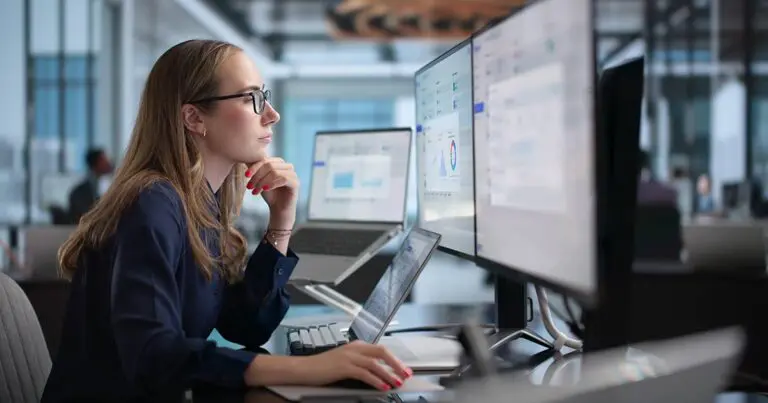 A woman wearing glasses works at a desk in a modern office, studying charts and data dashboards displayed across multiple computer monitors. She rests her chin on one hand while using a mouse, reflecting a focused, analytical work environment associated with technology integration roles.
