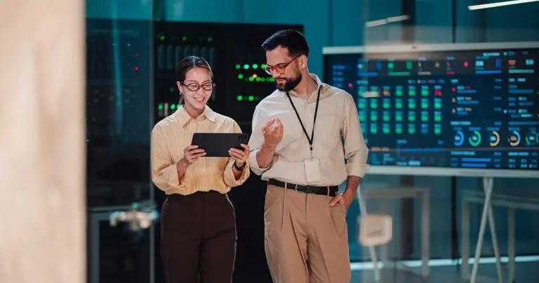 Two technology professionals wearing glasses review information on a tablet in a data center. Server racks and a large digital monitoring dashboard with colorful data visualizations fill the background, highlighting collaboration in digital transformation roles.