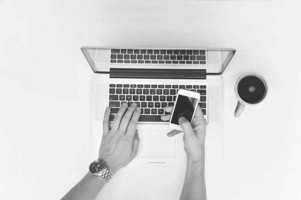 person working on a laptop on an outdoor table with a cup of coffee in one hand and smartphone in the other