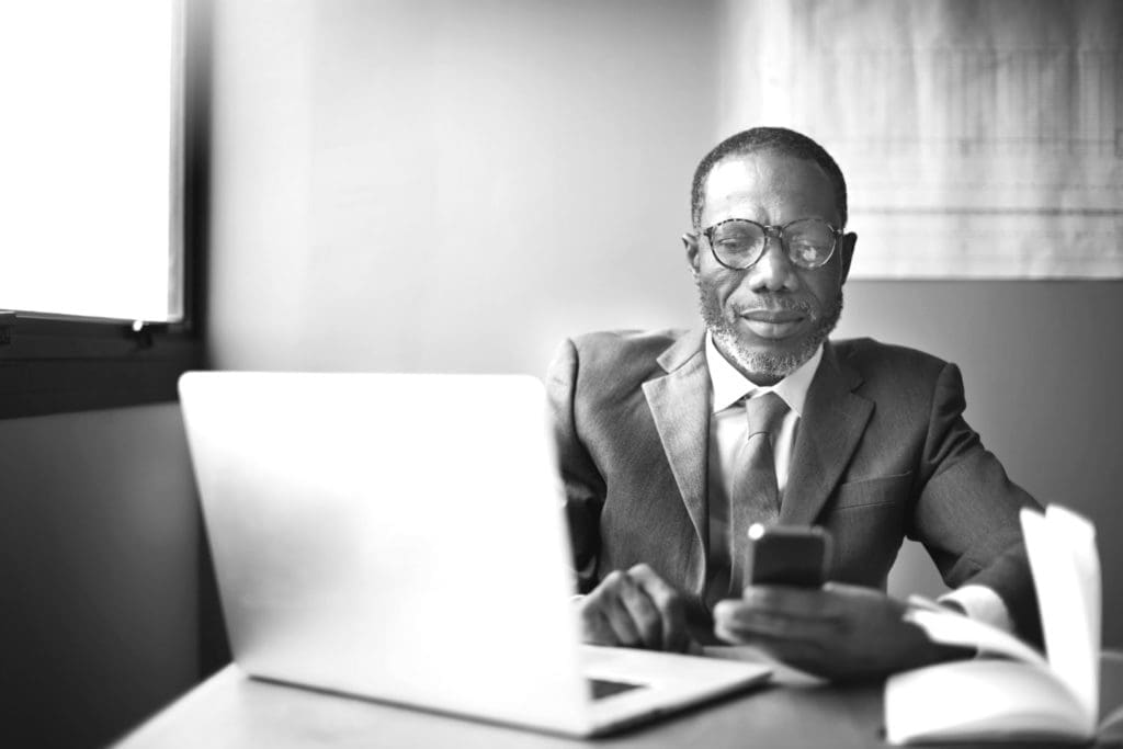 Person wearing a business suite in an office in front of a laptop looking at smartphone