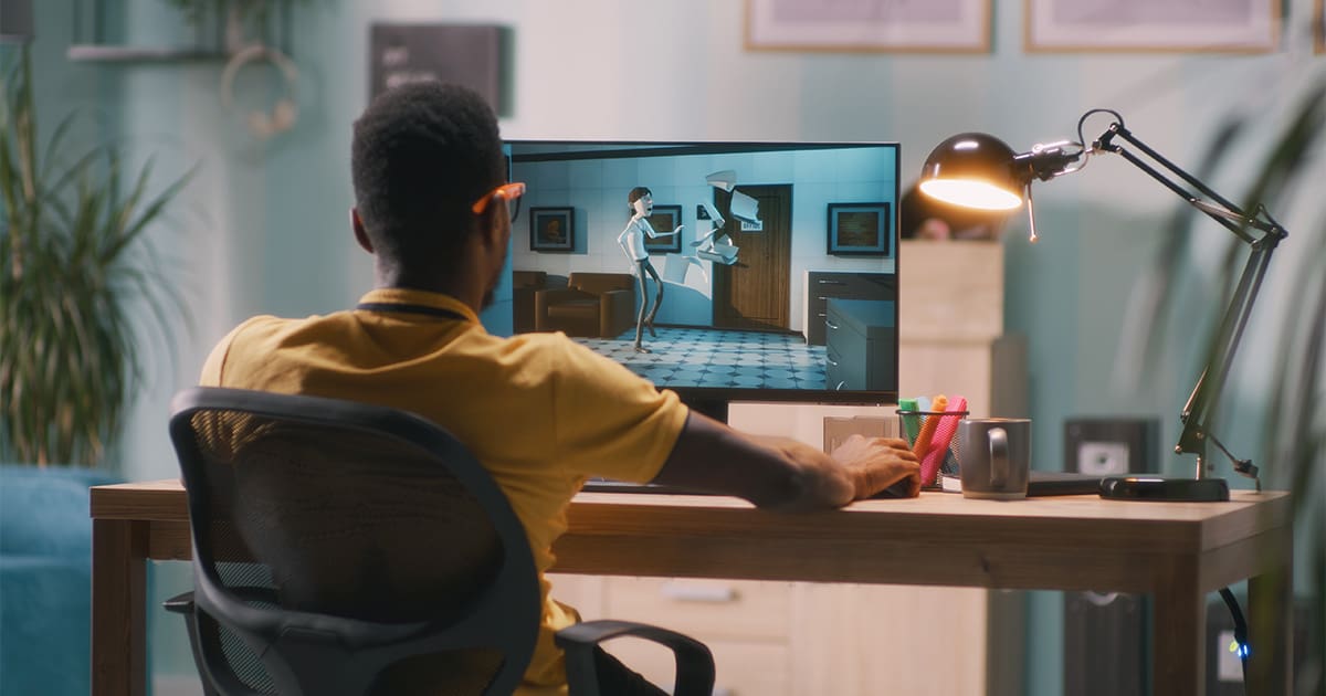 Black man in a yellow tshirt sitting at a desk working on a computer with his back facing the camera