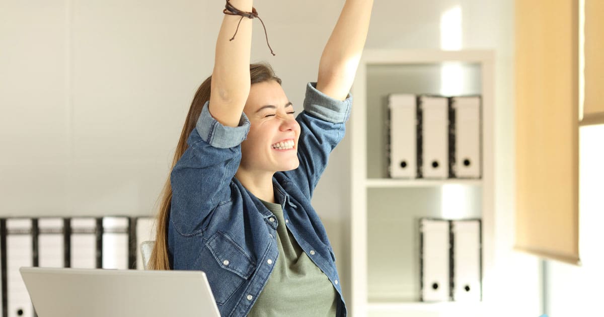 photo of a young brunette woman sitting in an office setting with a computer and raising her arms above her head triumphantly