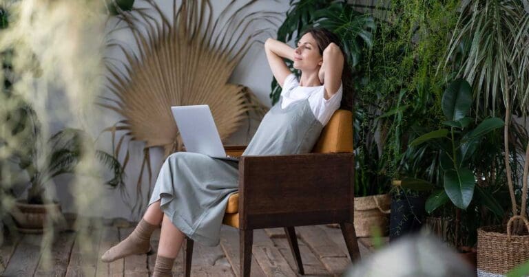 Photo of a woman sitting in a chair with a laptop in her lap and her arms behind her head leading back in a serene room with plants to signify work-life balance