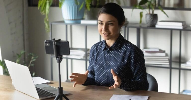Female with brown hair pulled back sitting at a desk speaking on a video conference call to a phone on a tripod