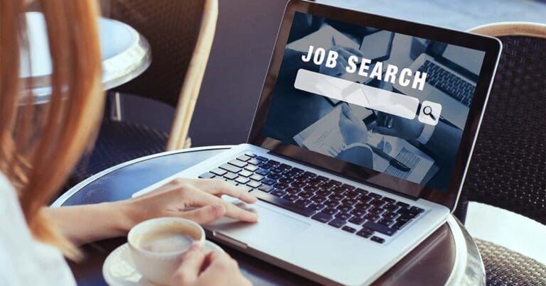 A photo of a laptop computer with the words job search over a search bar with a persons left hand typing while the right hand holds a cappuccino