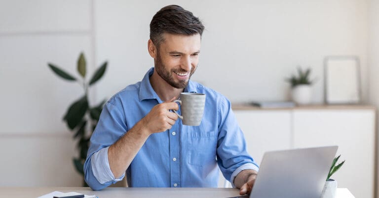 Photo of a white man with brown hair, a mustache, and beard wearing a blue button up collared shirt smiling and holing a cup of coffee while looking at a laptop in front of him